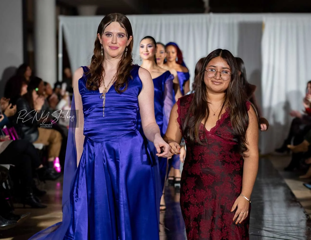 Two women walking down a runway at a fashion show, surrounded by seated audience members.