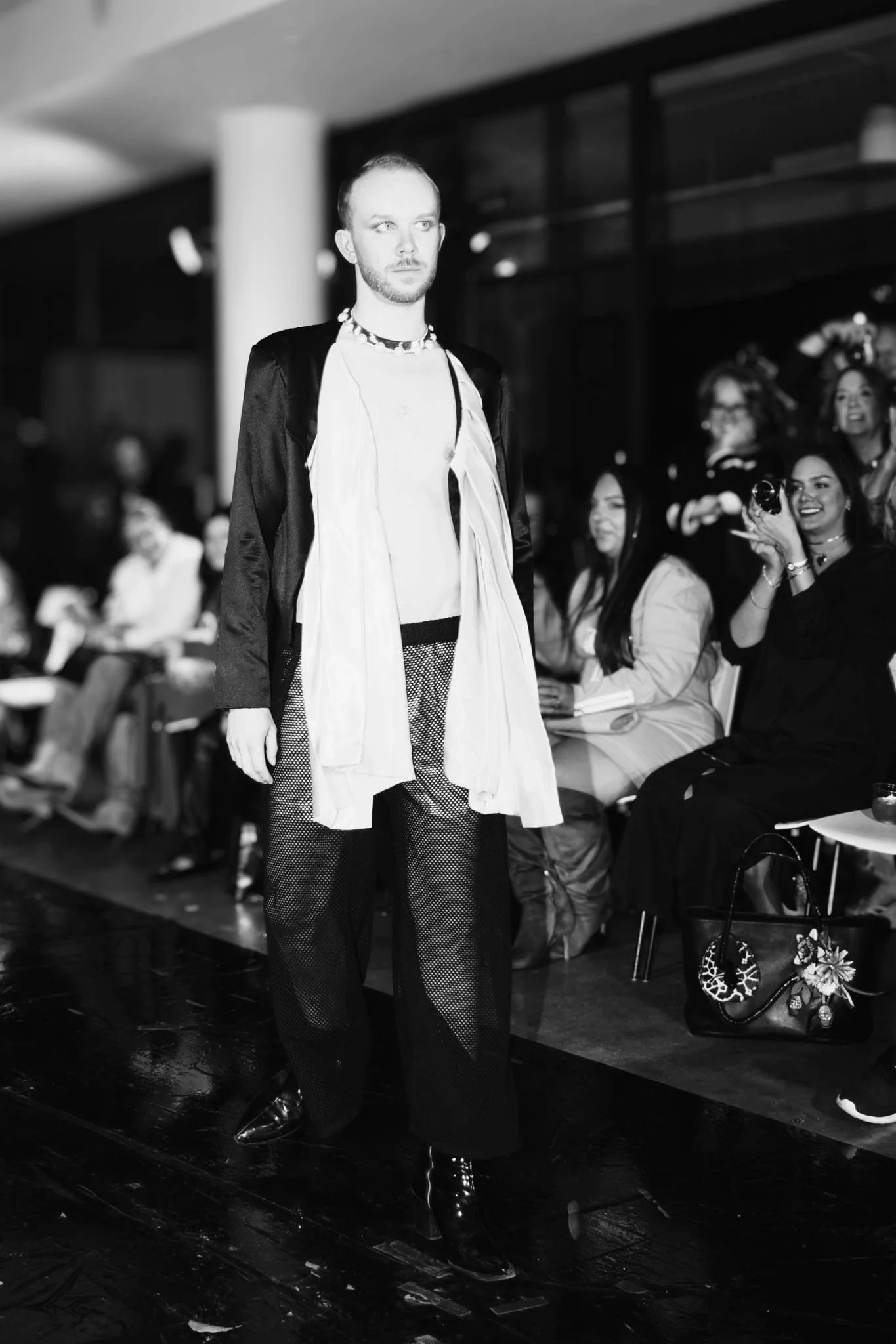 A male model on a runway at a fashion event, dressed in layered clothing with mesh pants, accessories, and shiny shoes, while a seated audience, including women smiling and holding drinks, observes.