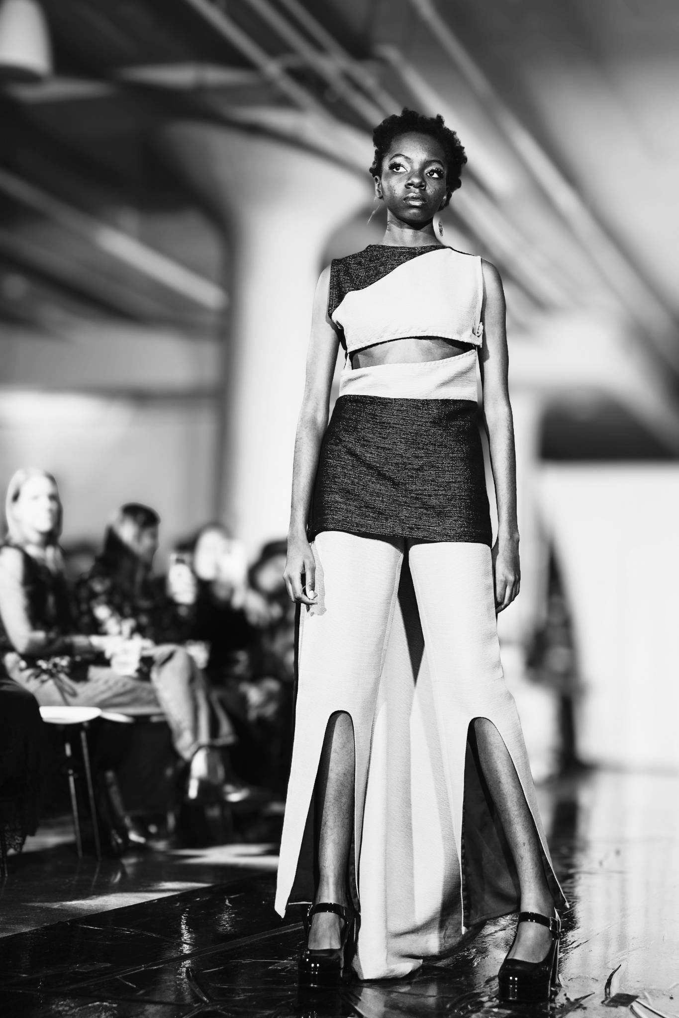 A woman walking on a runway during a fashion show, wearing bold, high-fashion clothing with wide-leg pants and a sleeveless top, with a serious expression, and audience members are visible in the background.