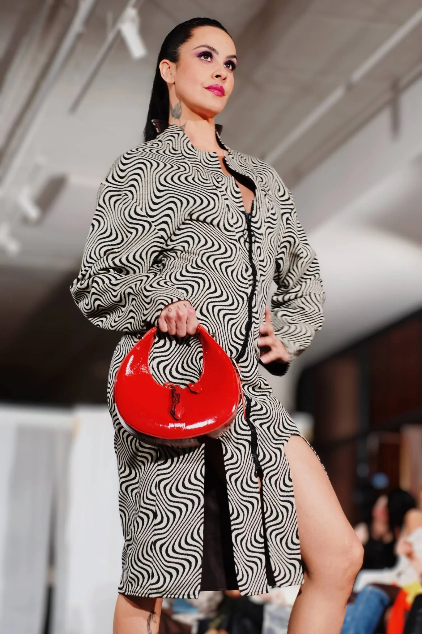 A woman with dark hair, wearing purple eye makeup and pink lipstick, stands confidently indoors. She is dressed in a black and white patterned outfit and holds a bright red handbag. The background shows a blurred indoor setting.