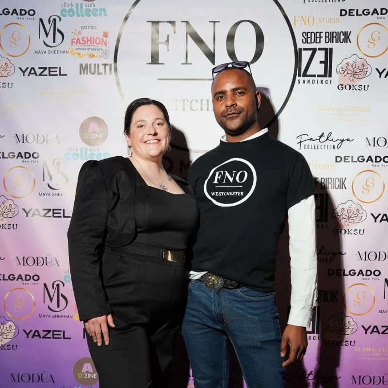 Two people standing together at a fashion event, with a backdrop featuring various brand logos and the large central circle logo reading 'FNO Westchester'.