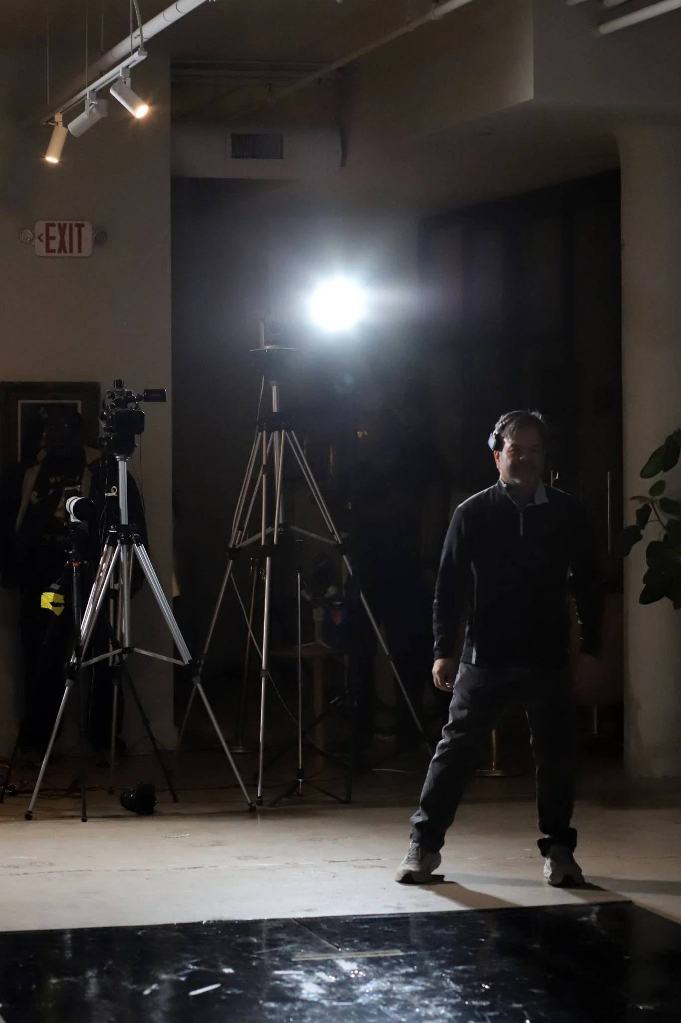 A man standing in front of a dark background with camera equipment and bright lights on tripods, in a dimly lit indoor space.