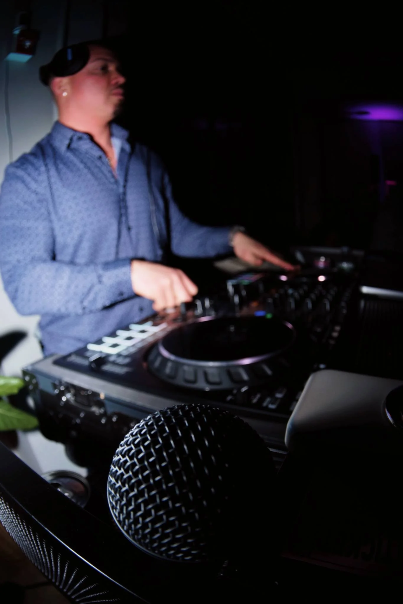 A person DJing at a dark indoor venue, wearing headphones and a patterned blue shirt, with DJ equipment and a microphone in the foreground.