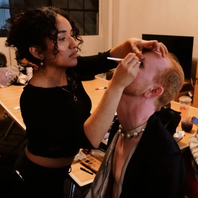 A woman with curly black hair uses a makeup brush to apply makeup to a person's lips. The person has short hair, a choker necklace, and is sitting calmly in front of a mirror in a room filled with makeup and beauty products.