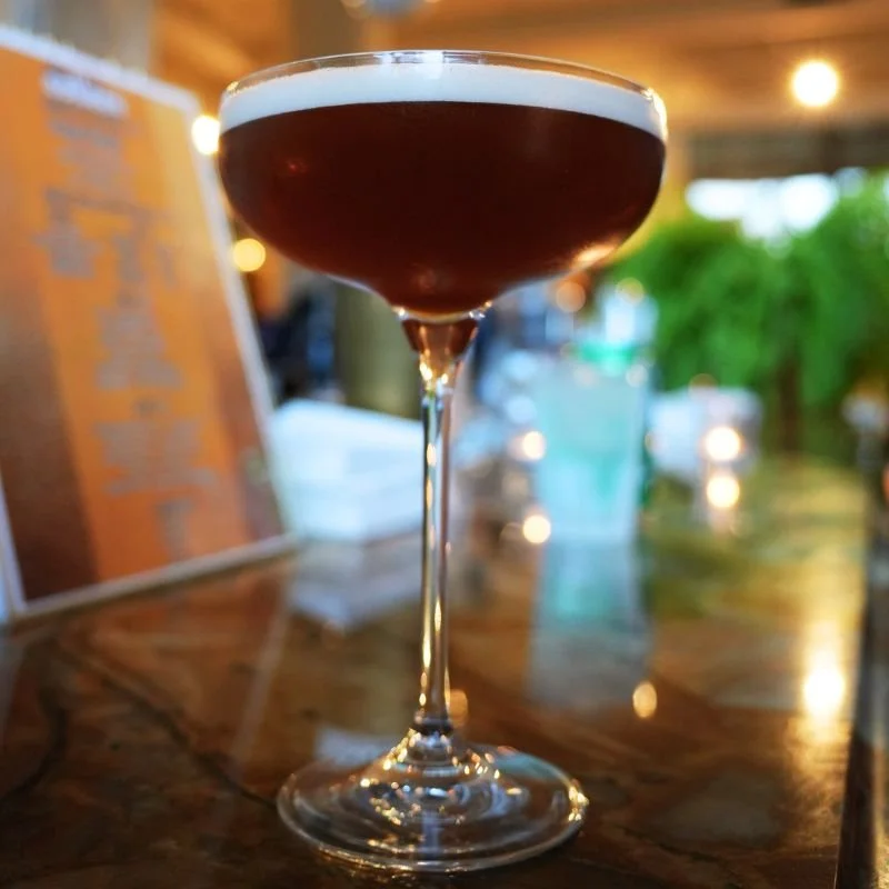 Close-up of a dark cocktail in a coupe glass on a wooden bar counter.