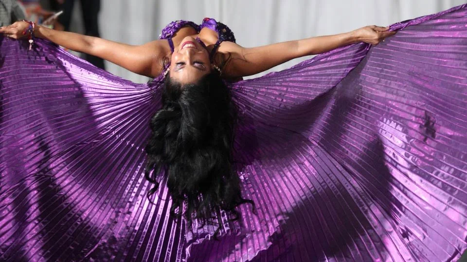 A woman in a vibrant purple dress holding the fabric outstretched, creating a dramatic and elegant pose, with an indoor backdrop.