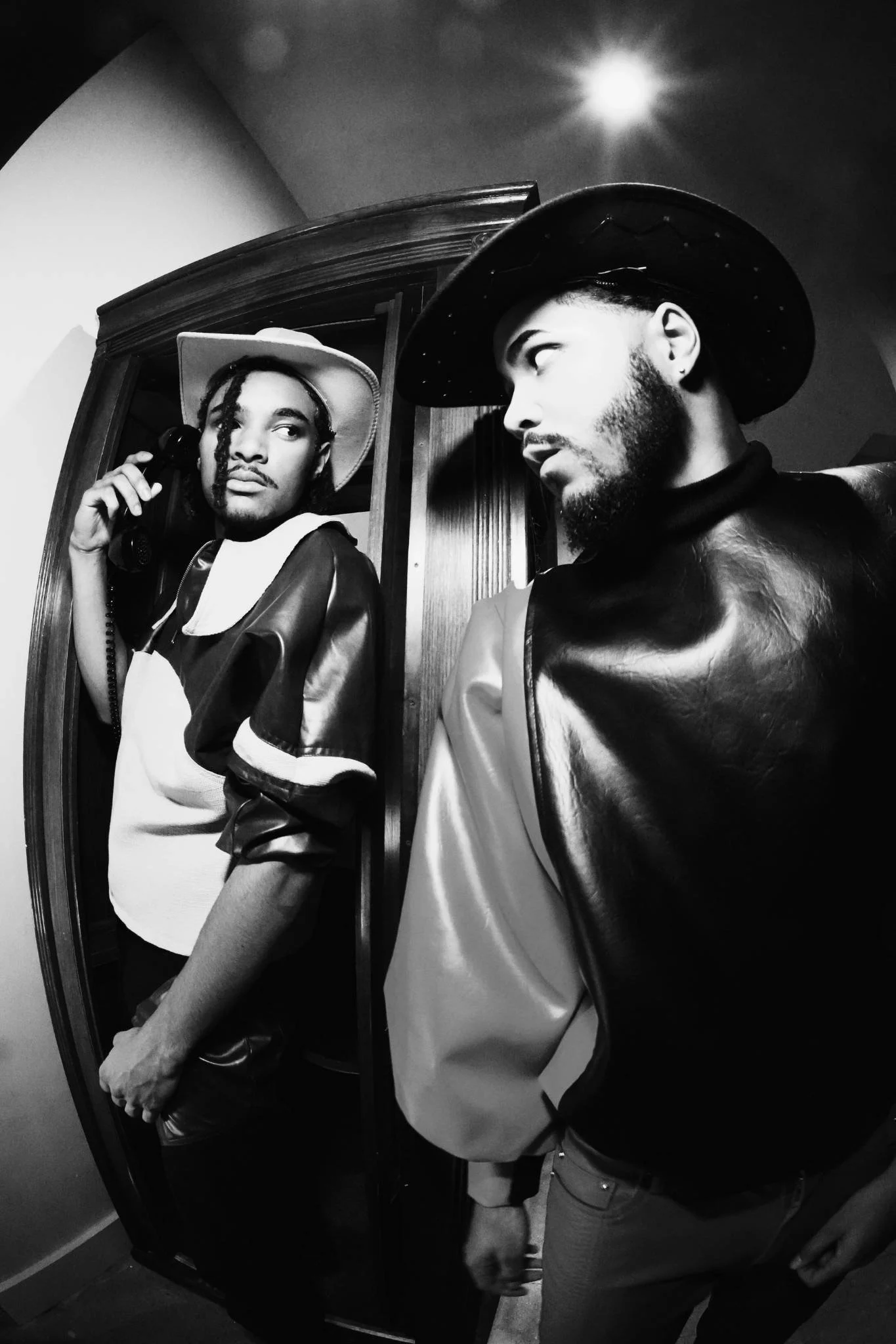 Two men with hats taking a selfie in front of a mirror in black and white. One man is holding a phone and looking at the other, who is wearing a cowboy hat and looking back.
