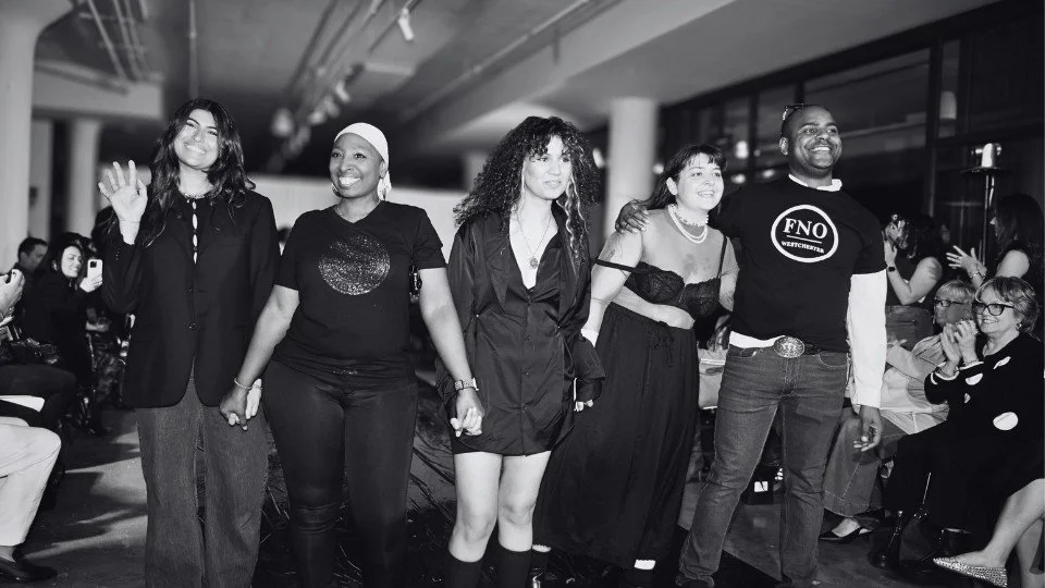 Group of five diverse people holding hands in a line at an indoor event, smiling and facing the camera, with seated audience and windows in the background.