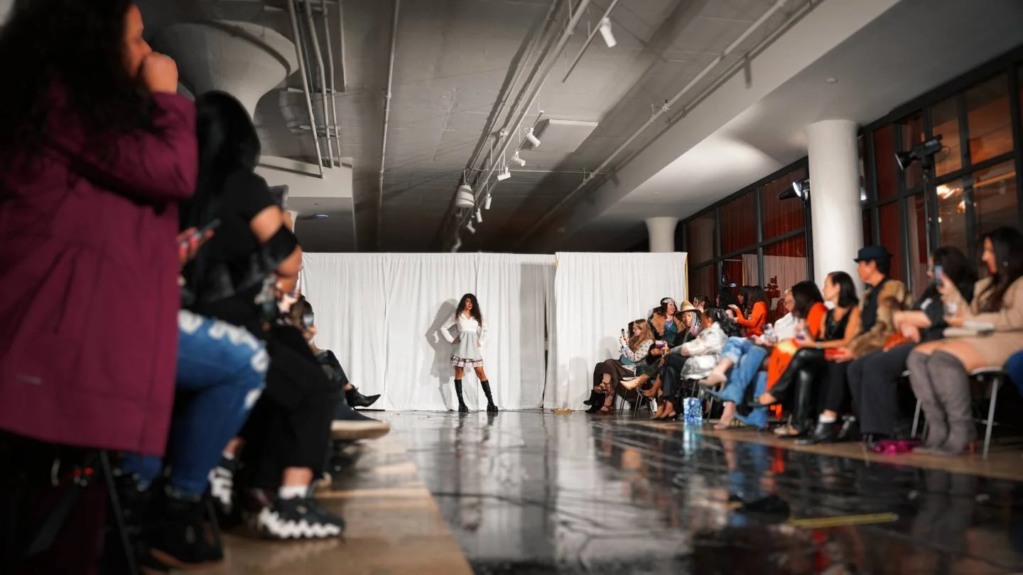 A fashion show runway with a model walking in front of a white curtain backdrop, surrounded by seated audience members on both sides of the runway, inside a modern venue with large windows.