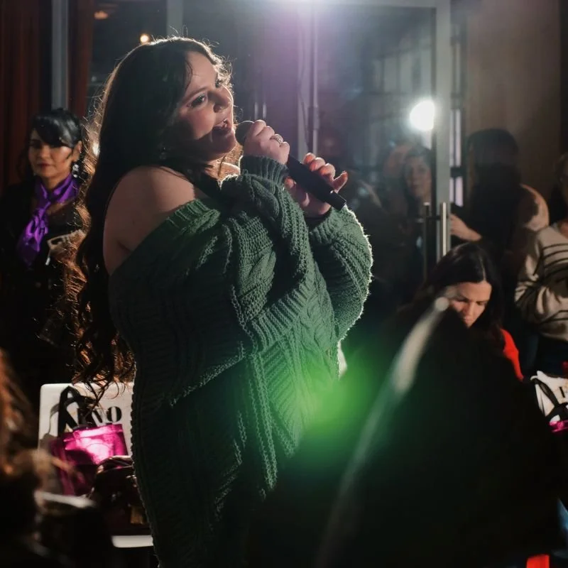 Young woman singing into a microphone at an indoor event with a crowd in the background.