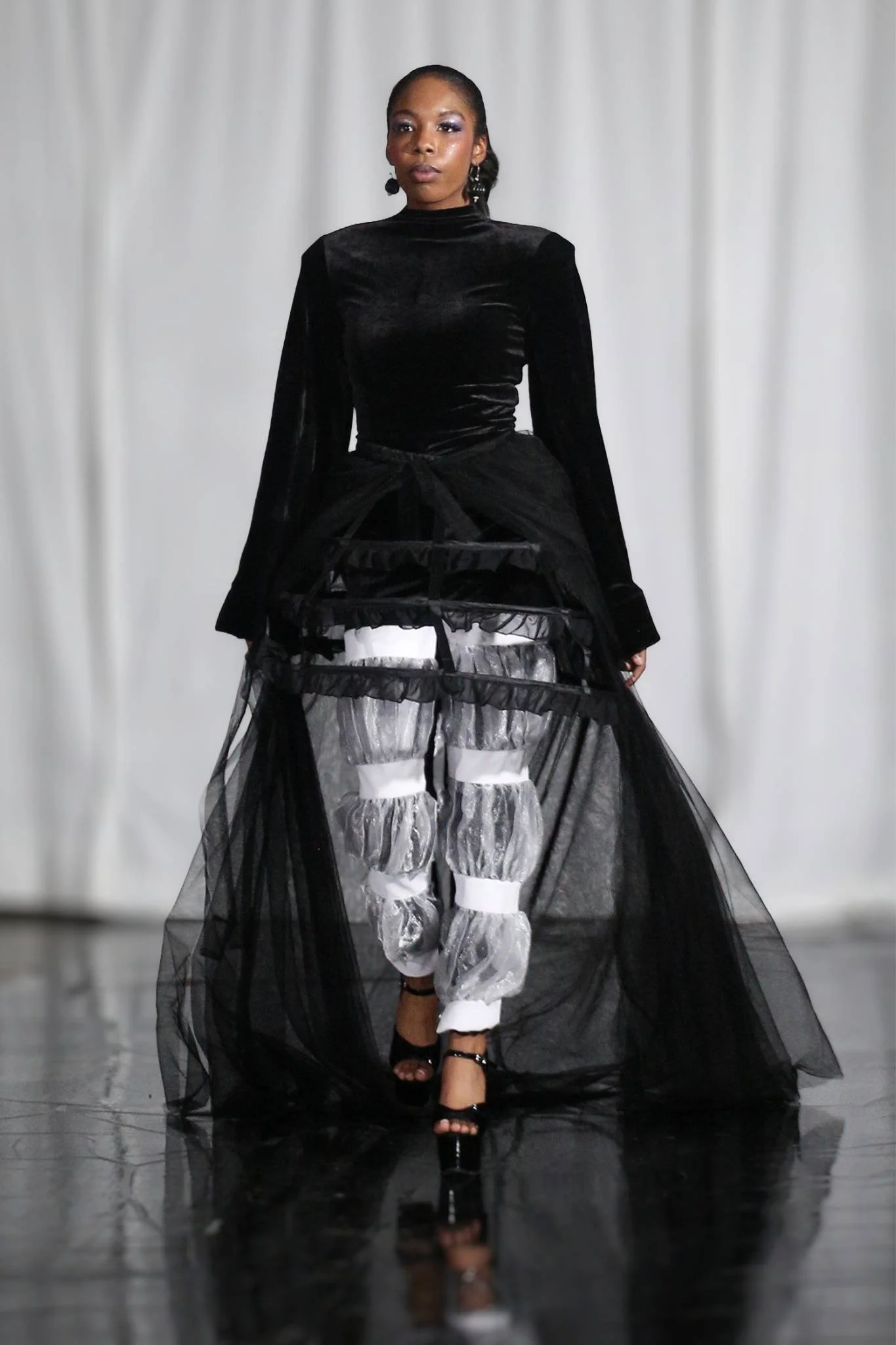 Model walking on runway wearing layered black and silver high fashion dress with sheer black overlay, dark hair in a bun, and black heels.