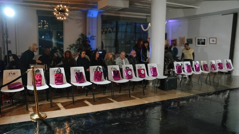 People sitting on chairs at an art gallery or event space, with pink gift bags on some of the chairs.