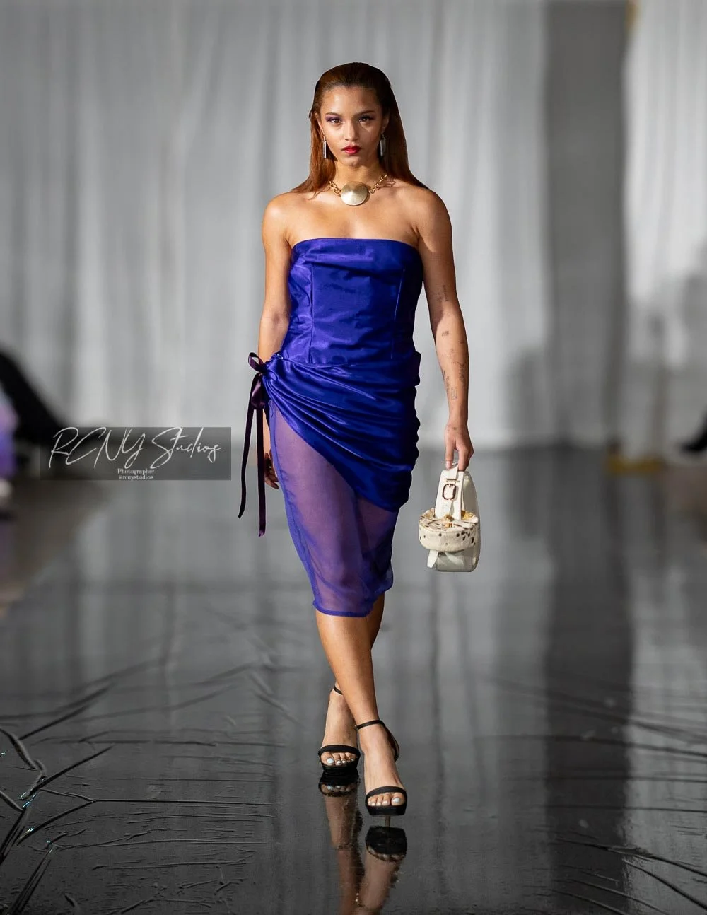 Woman walking on runway wearing a strapless blue dress with slit, black high heels, gold jewelry, and holding a small purse.