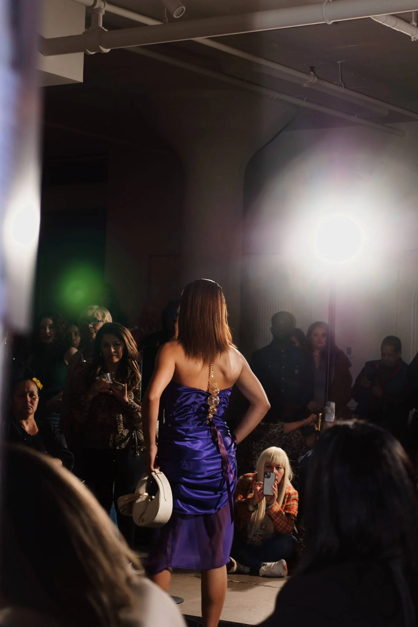 A woman in a purple dress with gold embellishments standing in front of an audience, with people seated and standing around in a dimly lit indoor venue, and a large bright light source on the right side of the image.