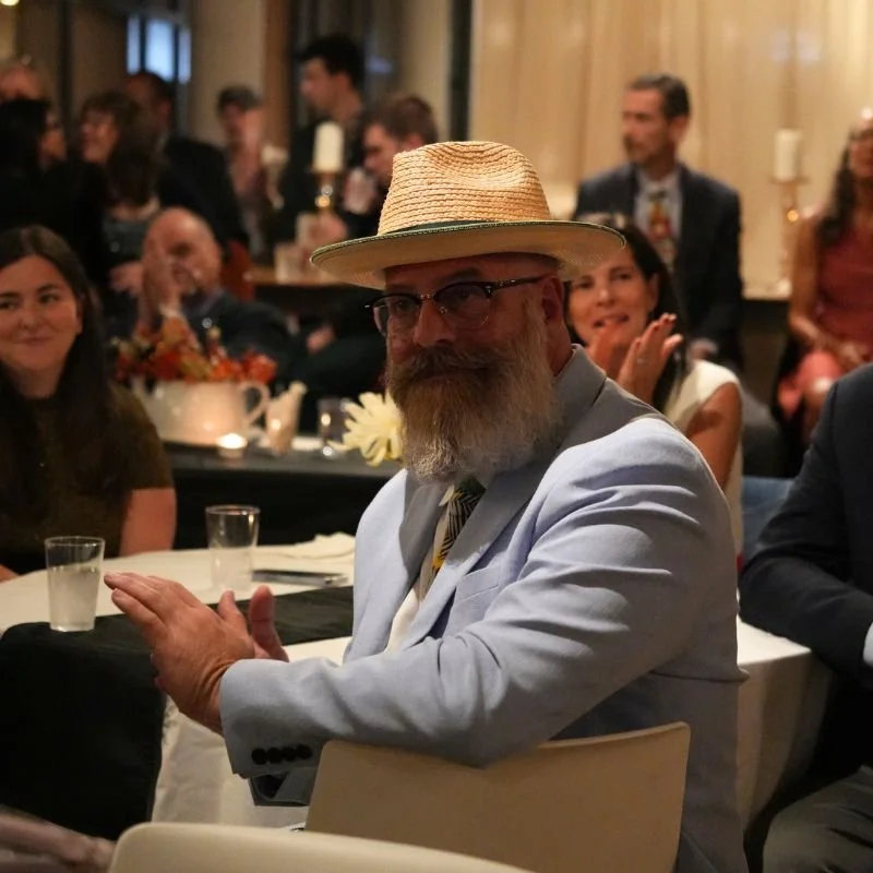 A man wearing a straw hat, glasses, a gray blazer, and a light-colored shirt sitting at a table during a social event, surrounded by other attendees who are clapping and smiling.