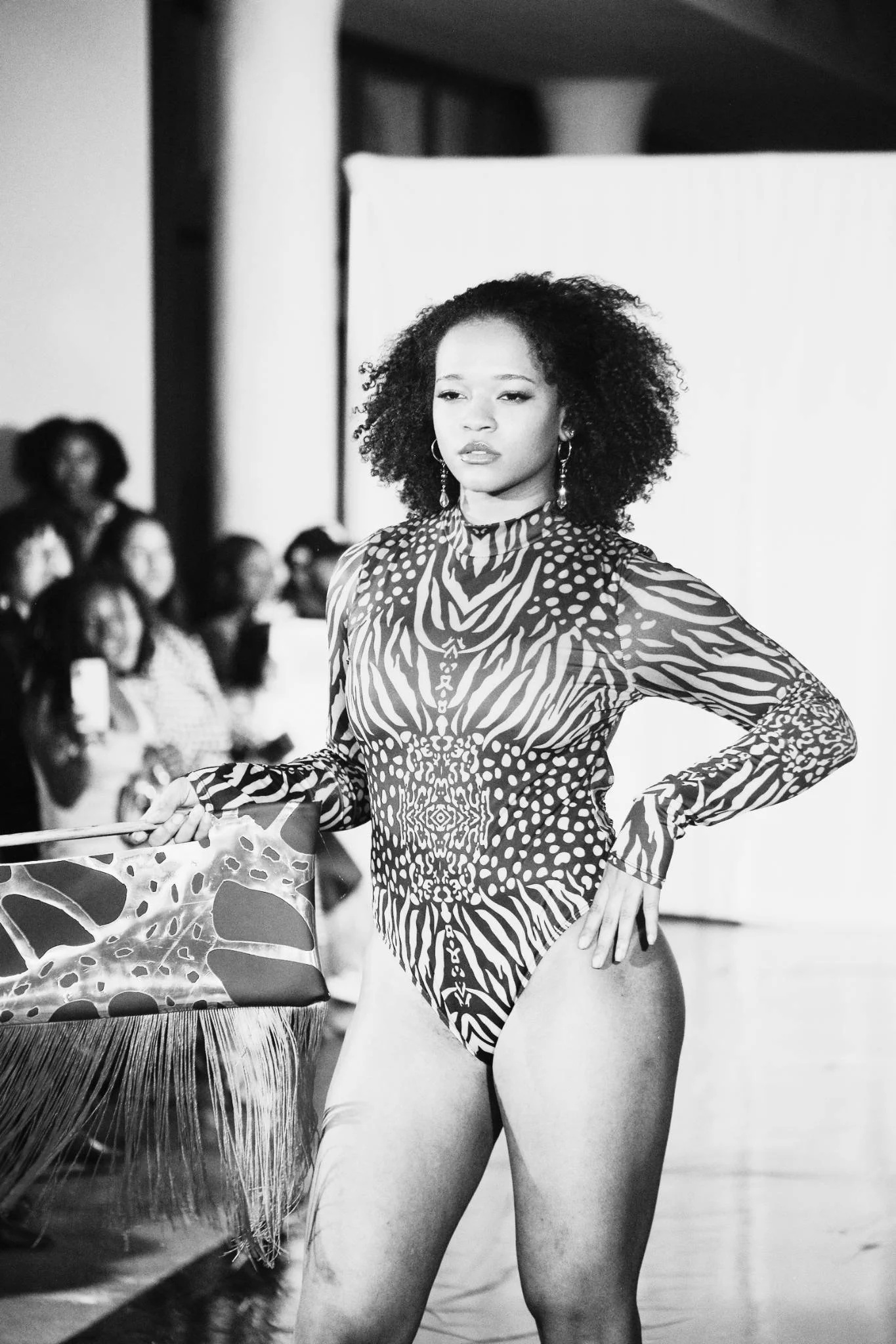 Black and white photo of a woman with curly hair in a patterned swimsuit standing with her hand on her hip, with an audience and a plain background visible behind her.