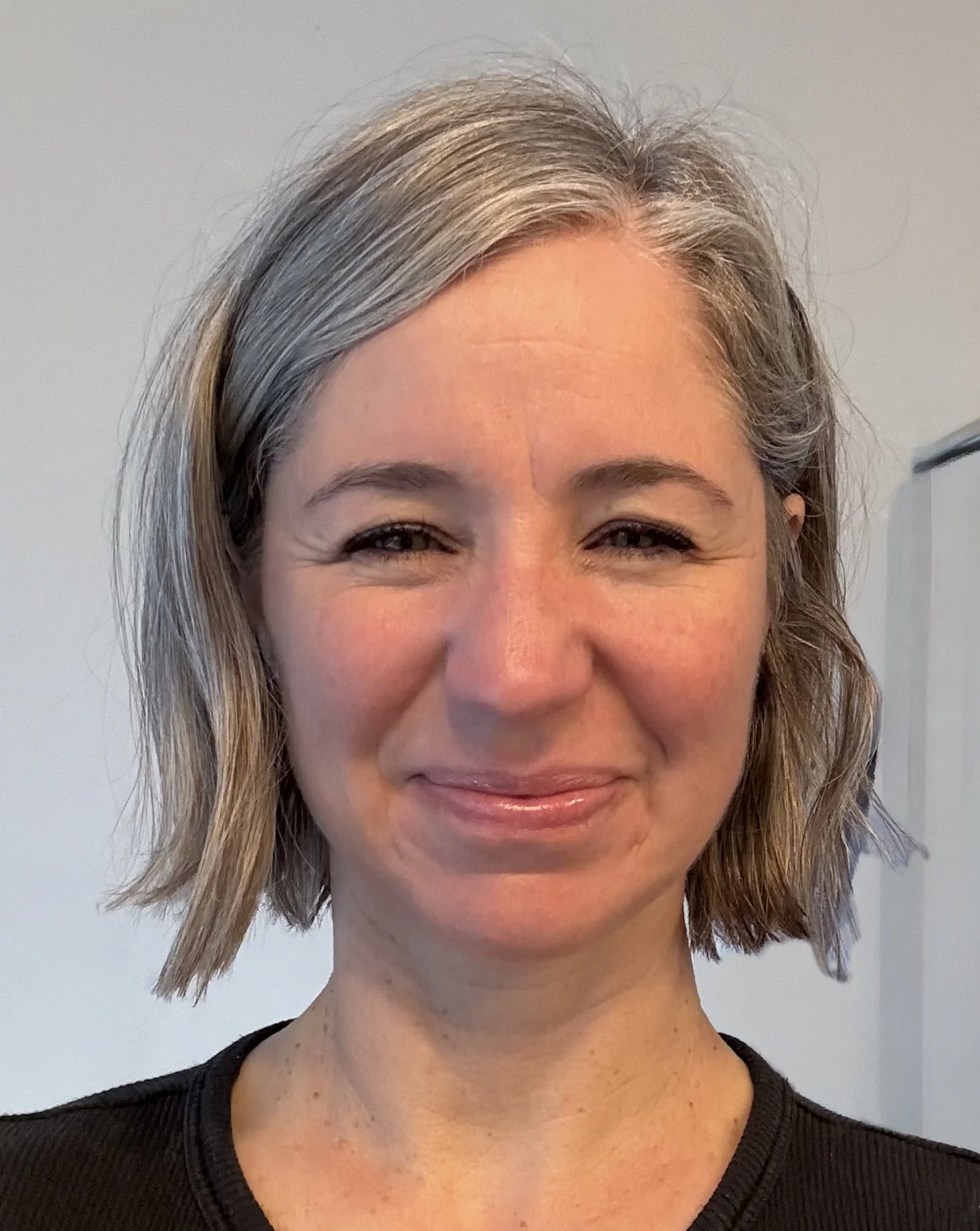 A woman with short, wavy gray hair smiling against a plain light gray wall, wearing a black top.