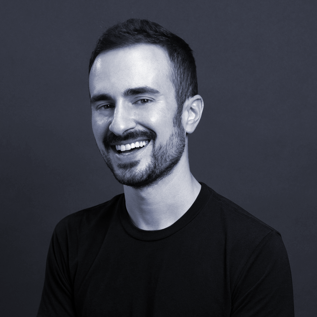 Smiling man with short dark hair, beard, wearing a black shirt, posed against a dark background.