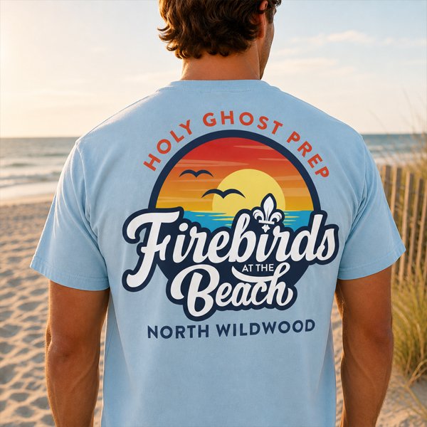 Person standing on the beach wearing a blue T-shirt with a sunset and wave graphic and text that reads "Holy Ghost Prep Firebirds at the Beach" and "North Wildwood."
