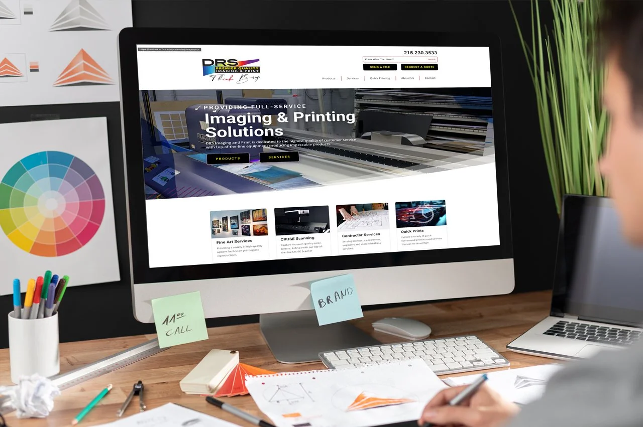 Desktop computer displaying the DRS printing and imaging website, surrounded by colorful design materials on a workspace desk.
