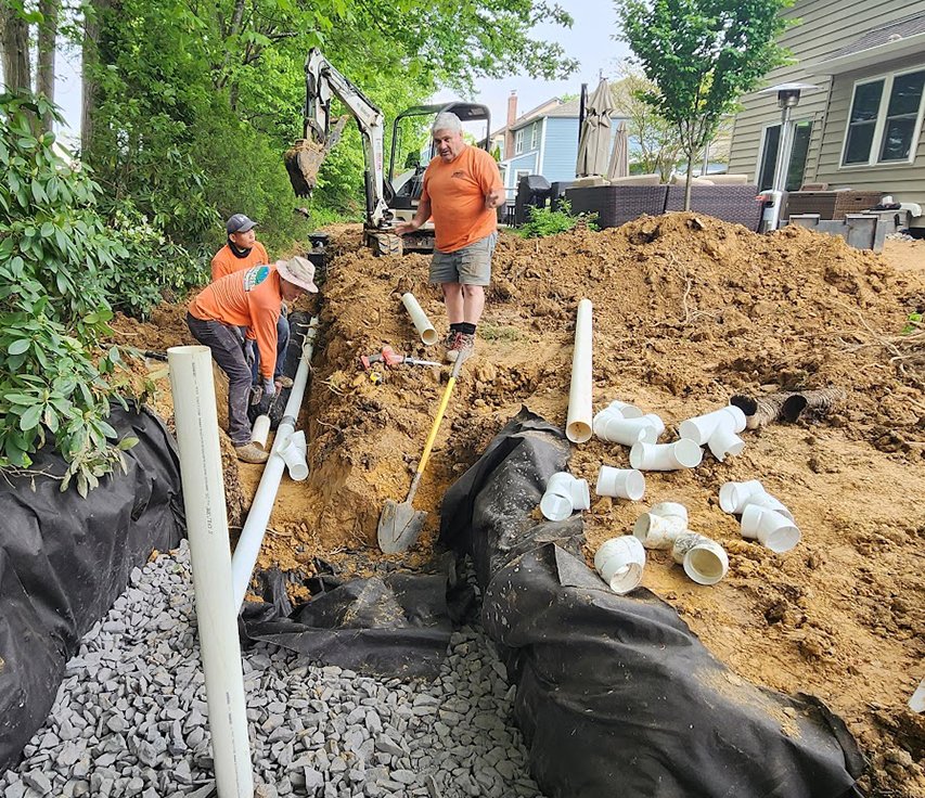 Three workers installing underground piping in a backyard with a small excavator, dirt piles, and scattered PVC pipes, with a house and trees in the background.