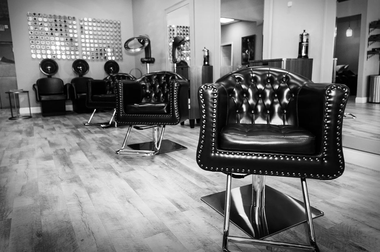 Black leather salon chairs with chrome bases, hairdryers, and salon equipment in a hair salon with wooden floors and modern decor, black and white photo.