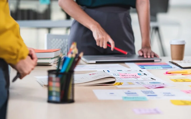 Two designers discussing project plans at a desk with notebooks, sticky notes, and branding color swatches.