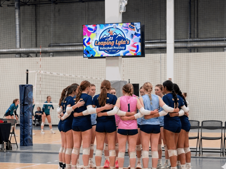 A group of young female volleyball players in a huddle on an indoor court, with a scoreboard and a sign that reads 'Leaping Lyla's Volleyball ProShop' in the background.