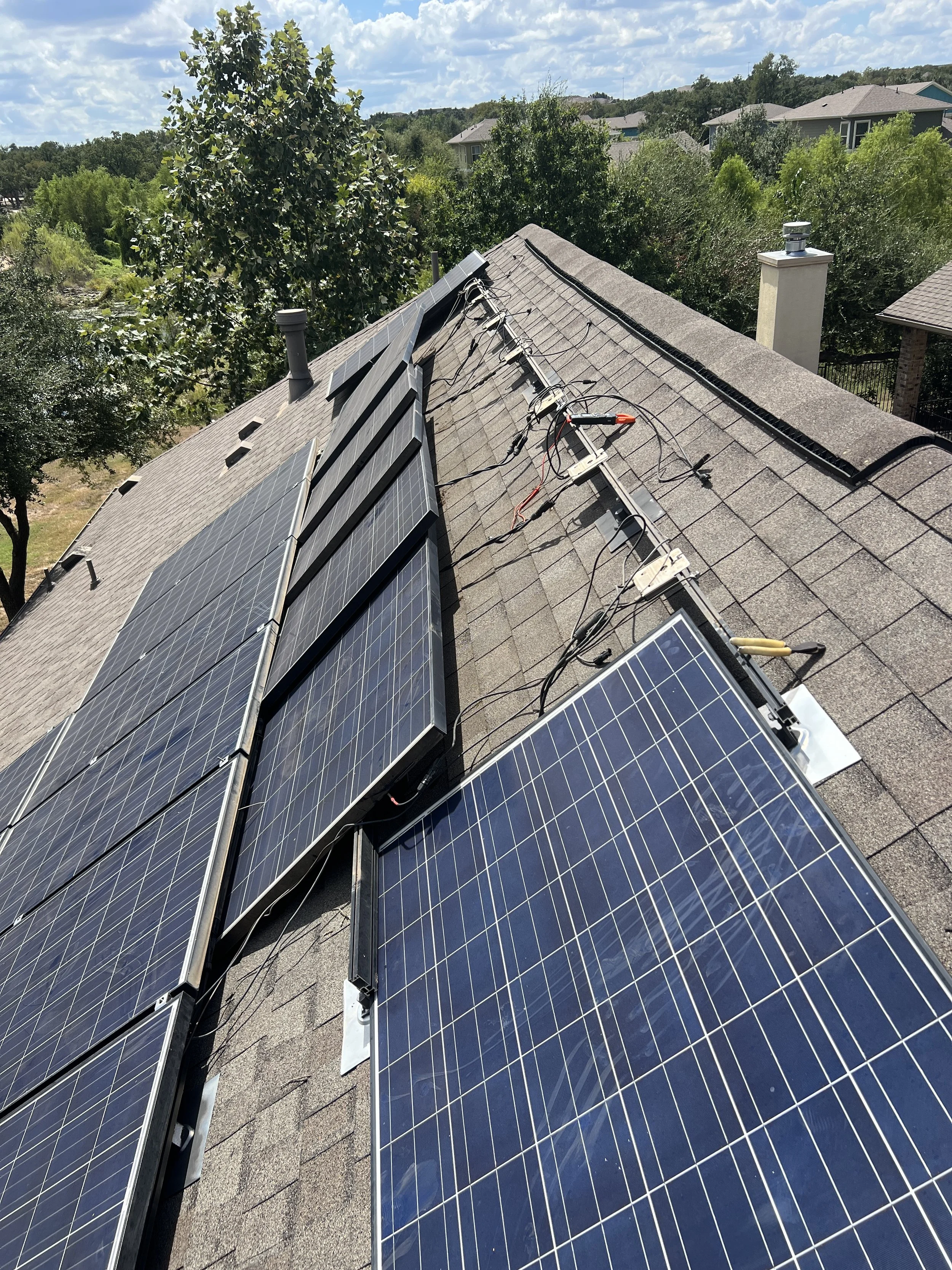Rooftop with installed solar panels, electrical wiring, and equipment, in a suburban area with trees and houses in the background under a partly cloudy sky.