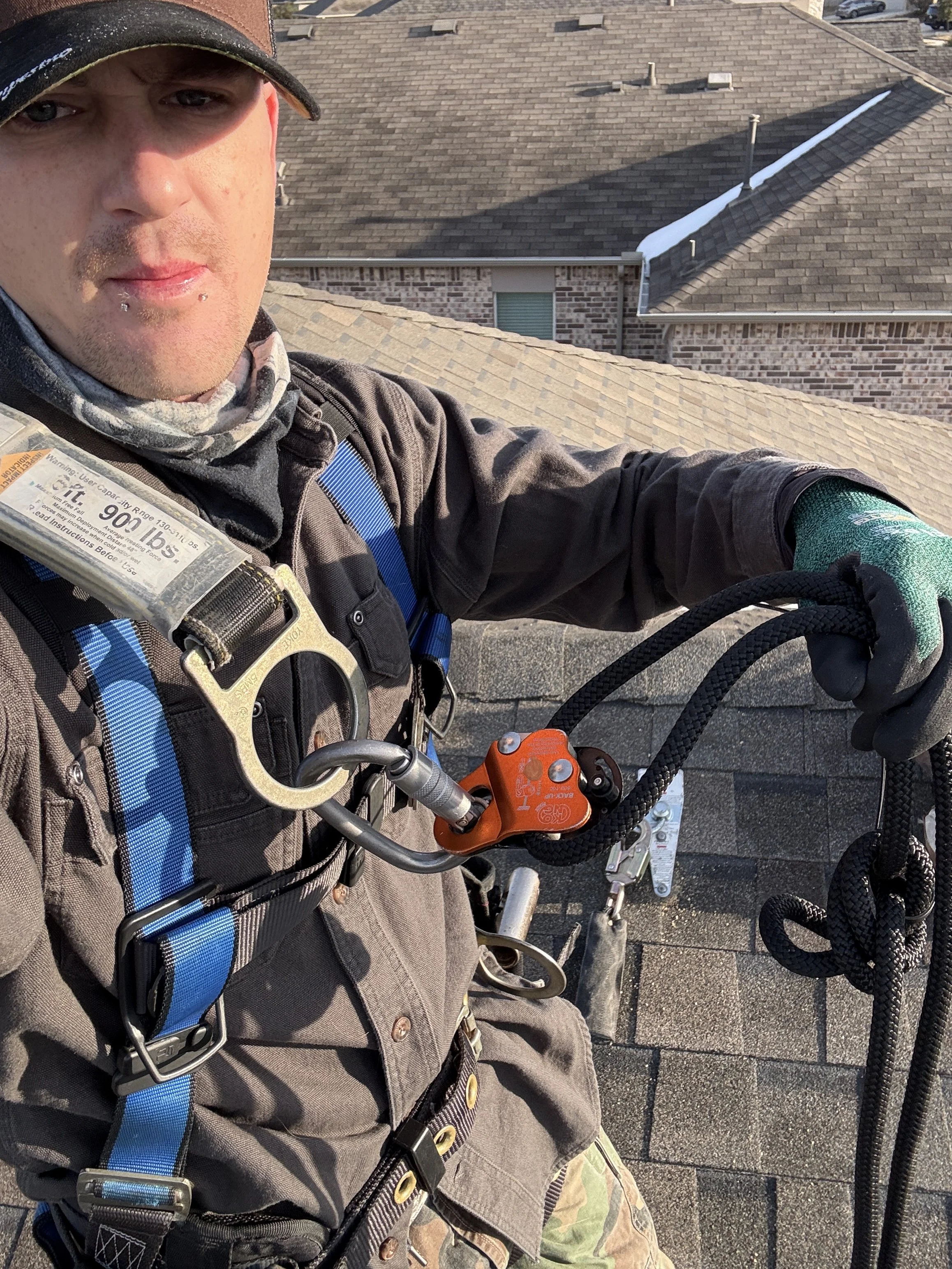 A worker wearing safety harness, gray jacket, and camouflage pants on a rooftop, with part of a chimney and a shingled roof in the background.
