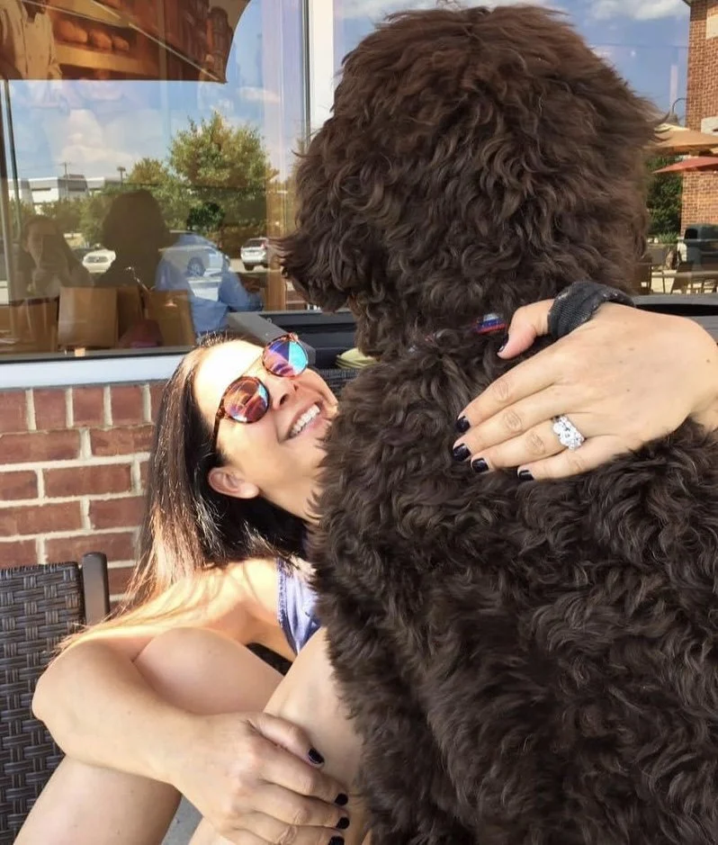 A woman in sunglasses smiling as she interacts with a large curly-haired dog, outside a cafe with a brick wall and glass window in the background.