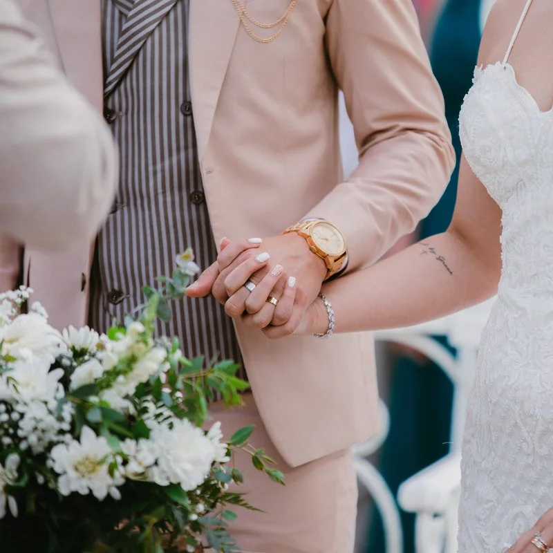 Close-up of a couple holding hands during a wedding ceremony, with wedding rings visible, a bouquet of white flowers, and the bride in a lace wedding dress.