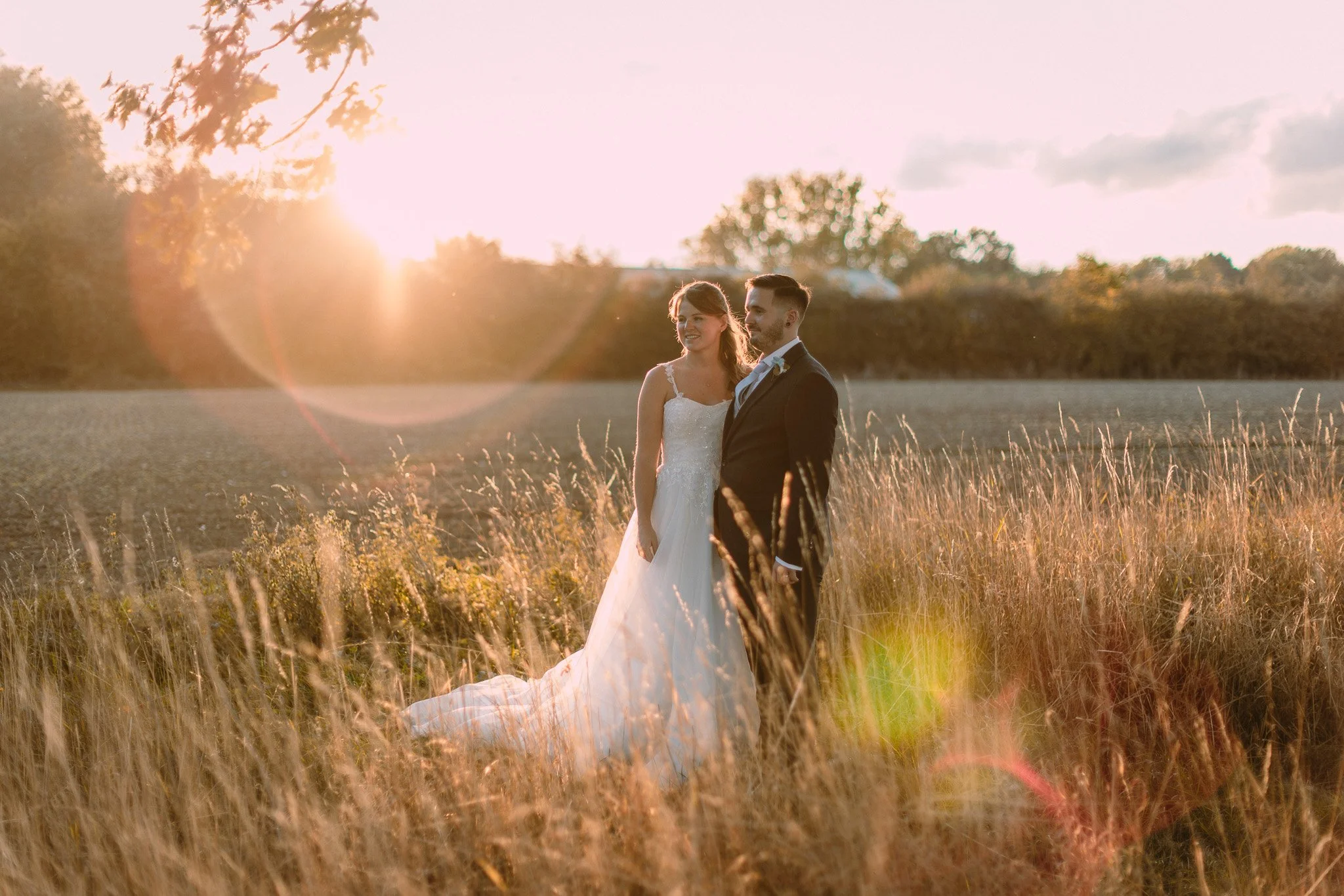A bride and groom standing in a field during sunset, smiling as the sun sets behind them.