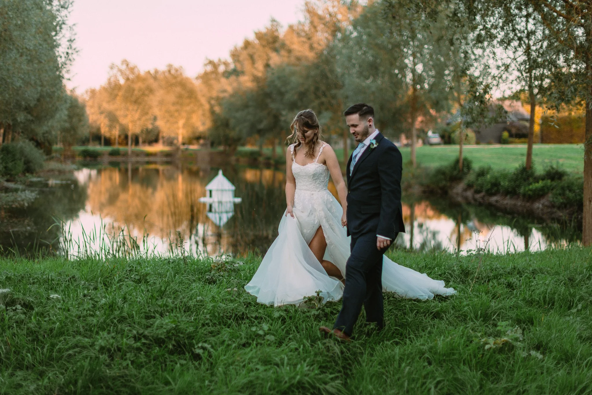 A newlywed couple walking along a grassy area by a pond during sunset. The bride is in a white wedding gown, and the groom is in a dark suit. The scene is surrounded by green trees and has a peaceful, picturesque setting.