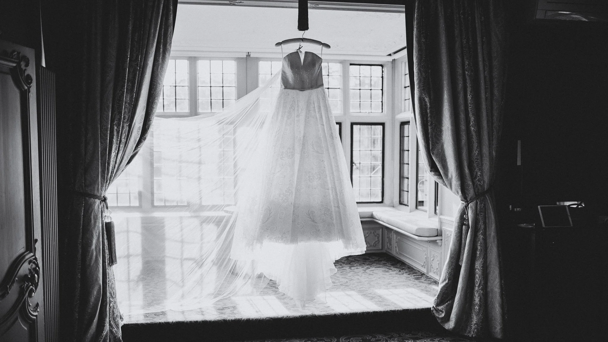 A wedding dress hanging in front of a large window with multiple panes, framed by heavy curtains on either side. The dress has a full skirt and a fitted bodice, with sunlight illuminating it.