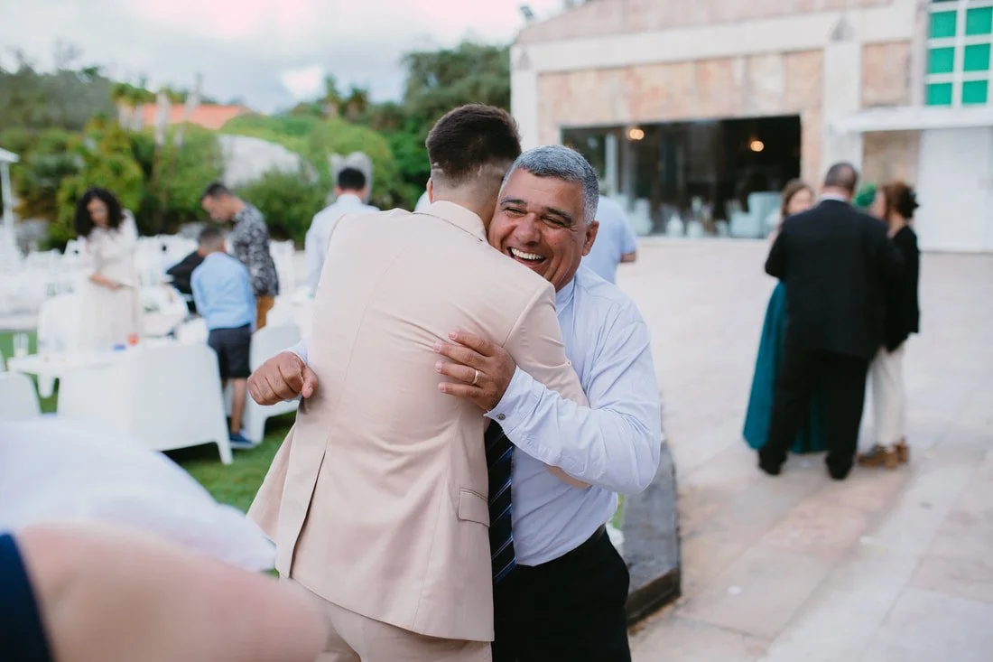 Two men hugging and smiling at an outdoor event with tables and other guests in the background.