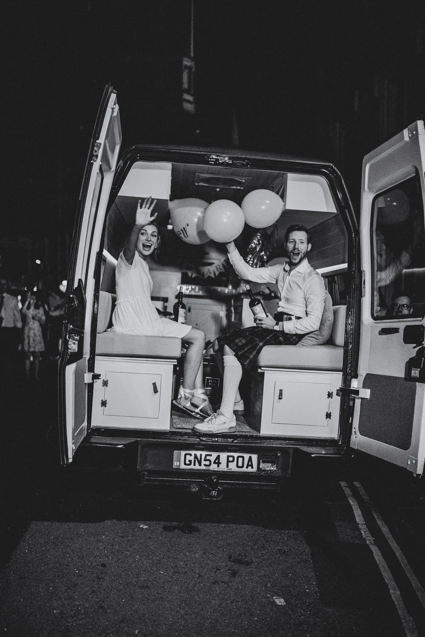 Two smiling people, a woman and a man, sitting in the back of a vehicle decorated with balloons during night time, waving and holding bottles.