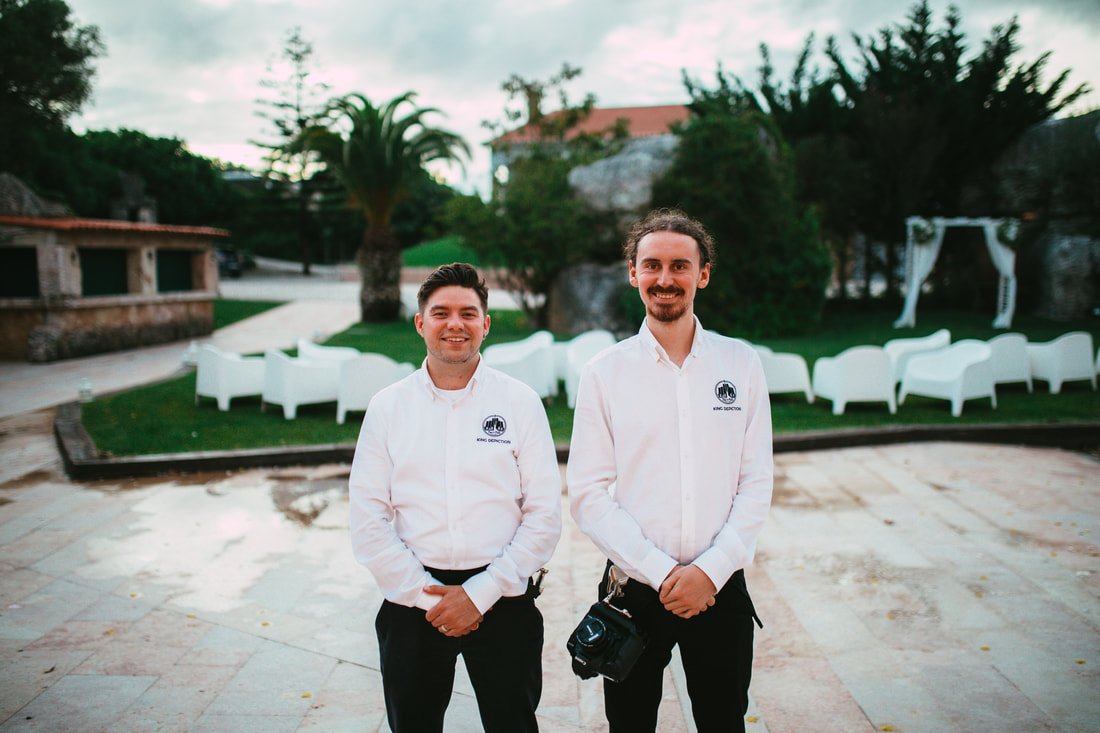 Two men standing outdoors in front of white chairs and greenery, both wearing white shirts with a logo, smiling at the camera.