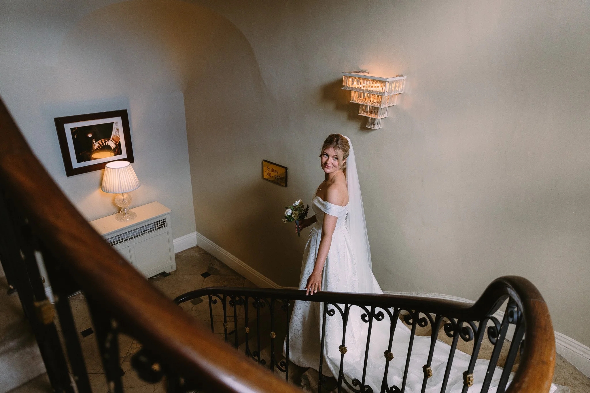 A bride in a white wedding dress and veil holding a bouquet of flowers, standing on a staircase landing with a painted wall and framed photo behind her. She is smiling at the camera.