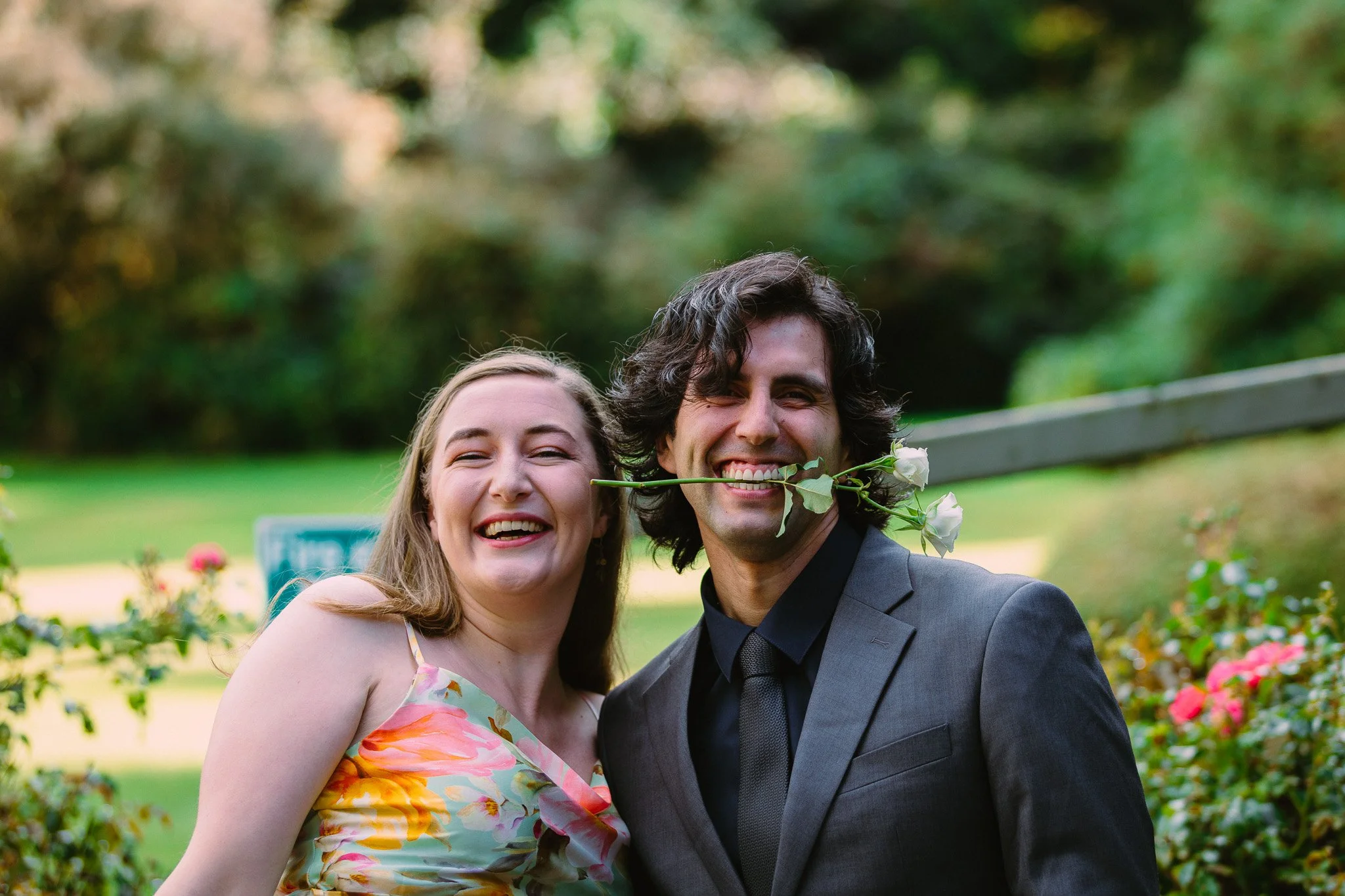 A smiling woman in a colorful dress with a floral pattern and a man in a dark suit are outdoors, with a background of trees and flowers.