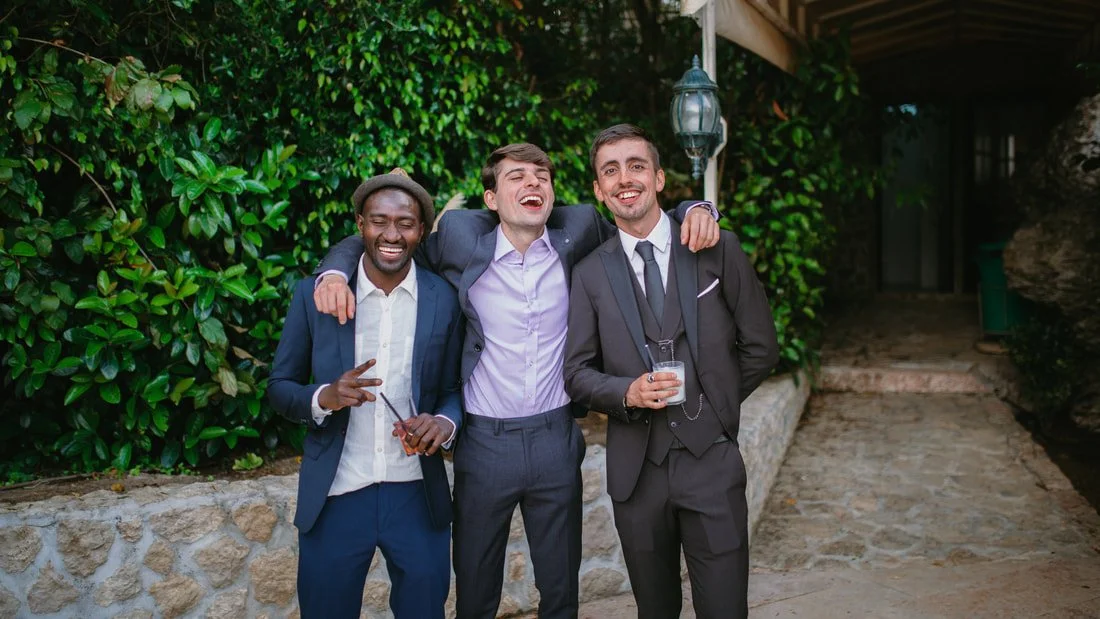 Three men in suits enjoying drinks and smiling together outdoors.