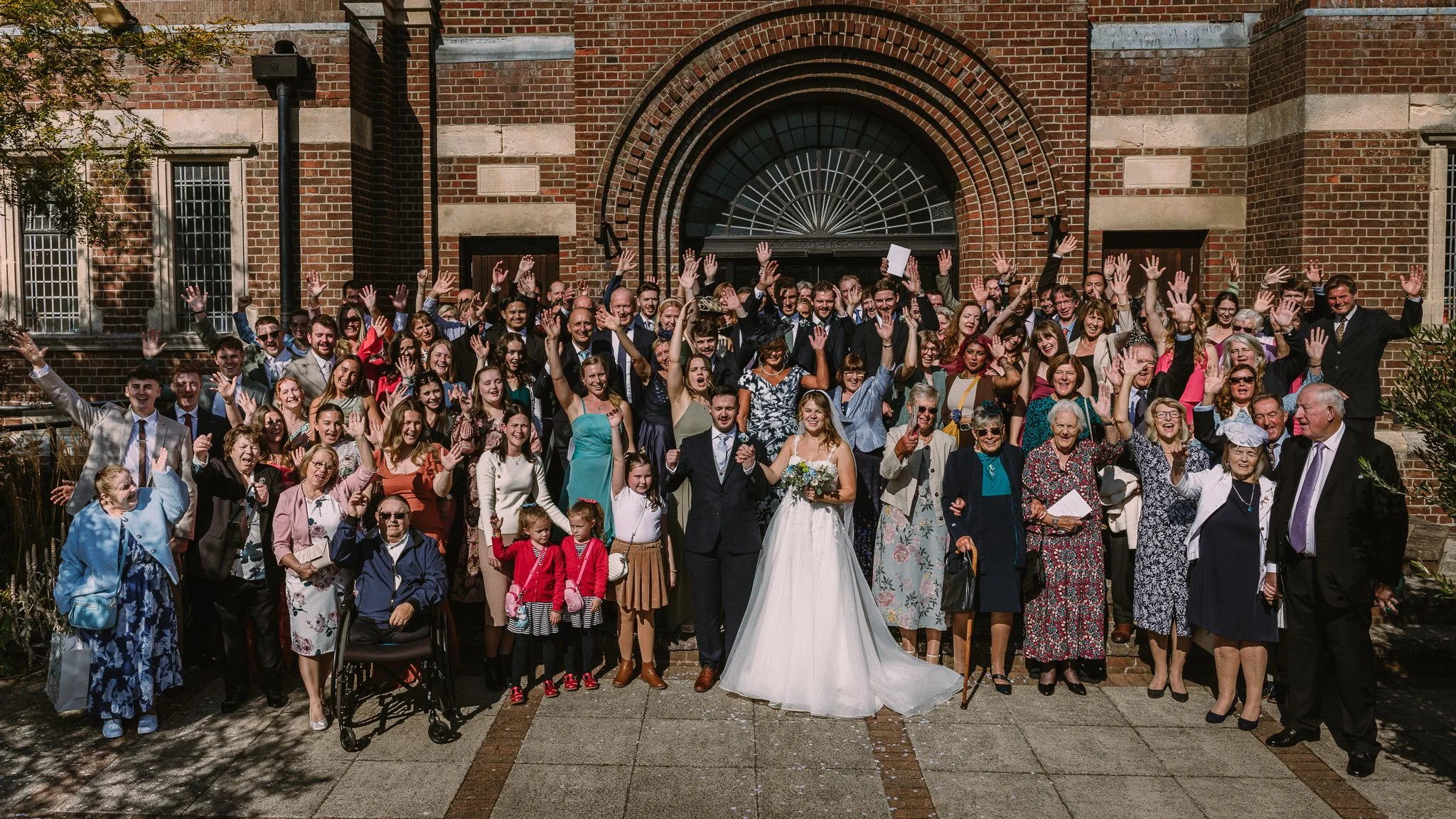 A large group of people gathered outside a brick building, celebrating a wedding. The bride and groom are in the center, waving with the crowd around them.