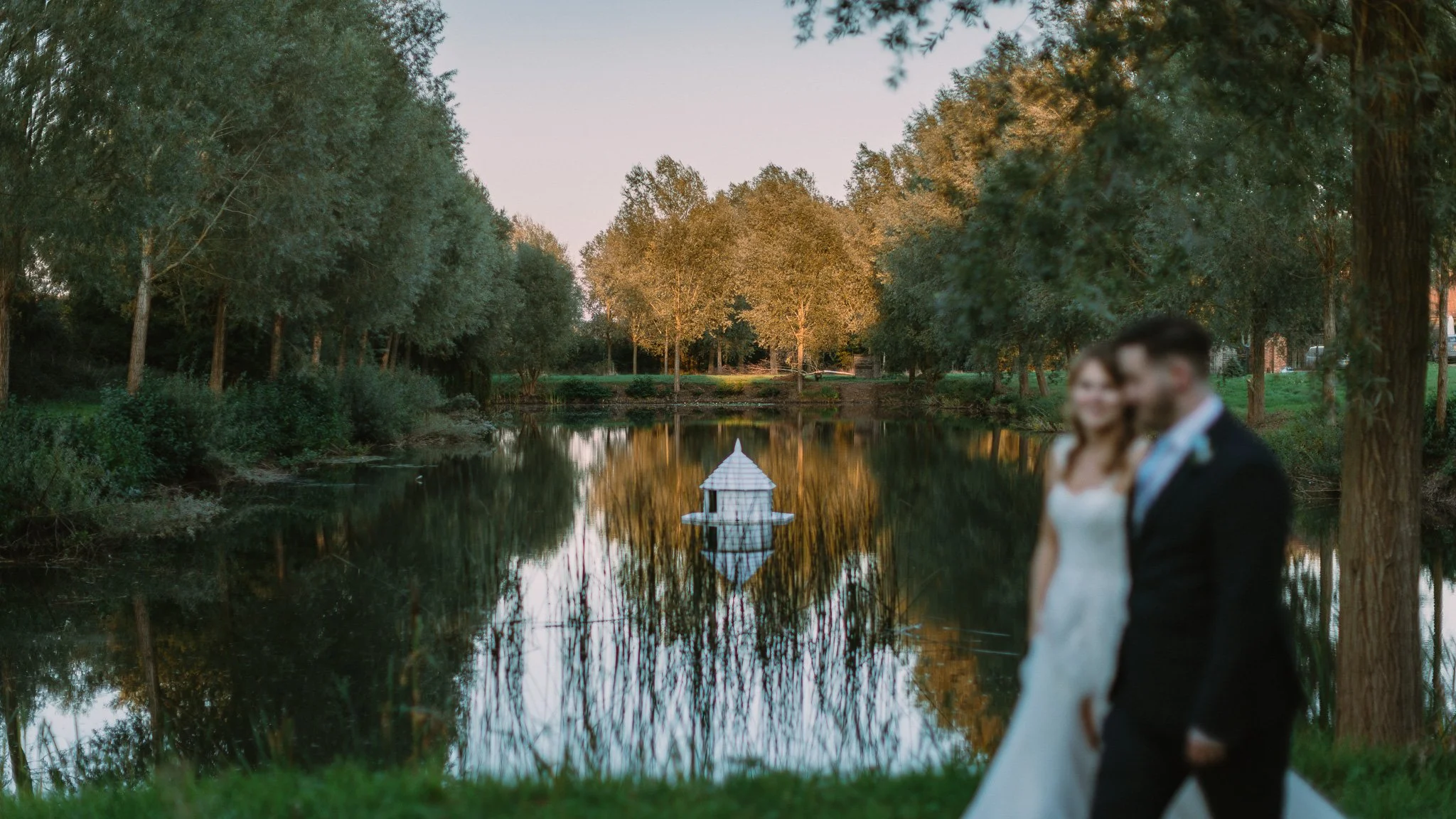 A couple in wedding attire standing by a pond in a wooded park with trees reflecting in the water and a small white structure on the pond.