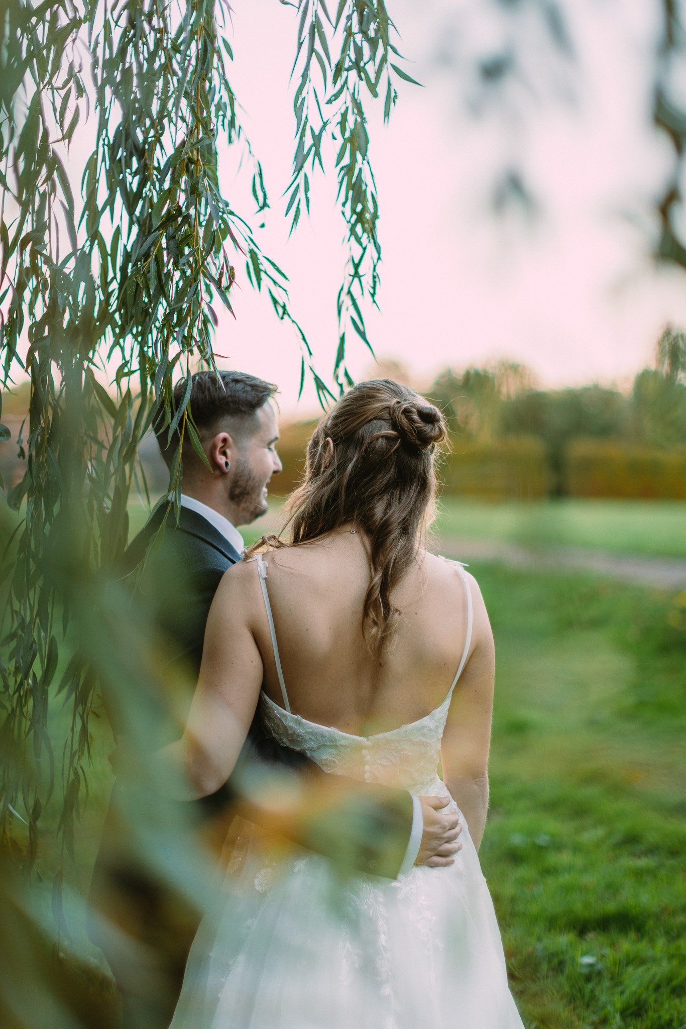 A bride and groom standing outdoors in a grassy area, with the bride wearing a white wedding dress with thin straps and the groom in a dark suit, partially obscured by hanging green leaves.