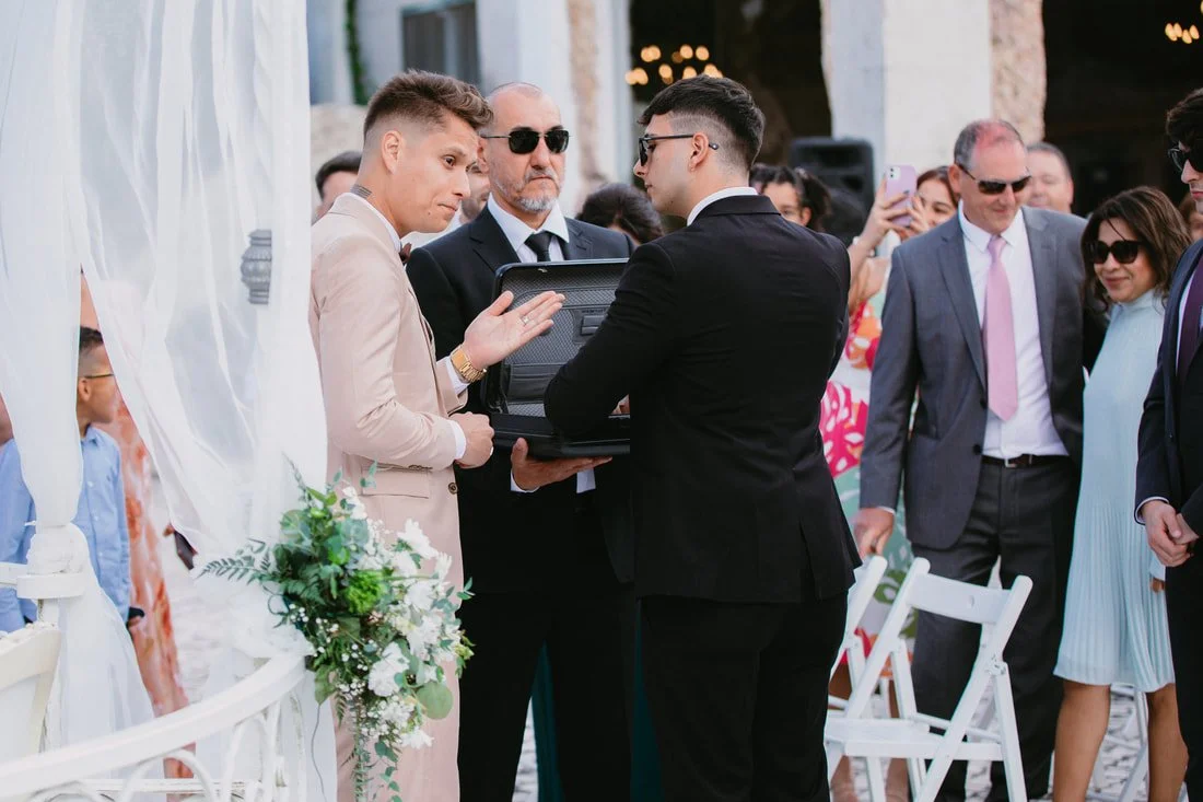 At a wedding ceremony, two men hold a small black box as they prepare to exchange rings. Guests watch and take photos, with a floral decoration and white chairs in the background.