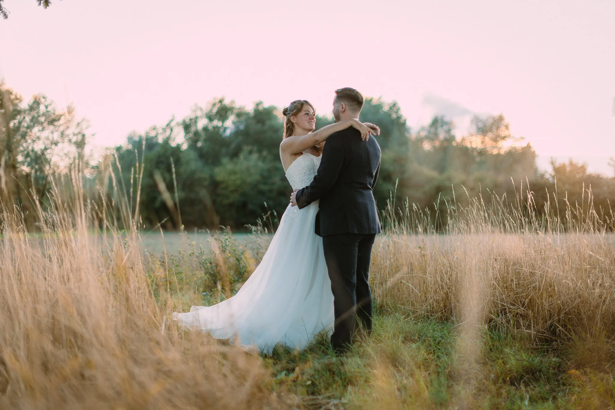A bride and groom in wedding attire dancing in a field of tall grass with trees in the background during sunset.