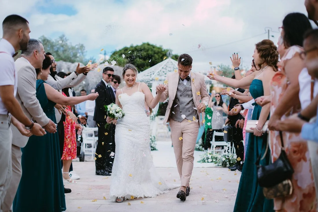 A bride and groom walking hand in hand down an outdoor aisle while friends and family throw flower petals celebrating their wedding.