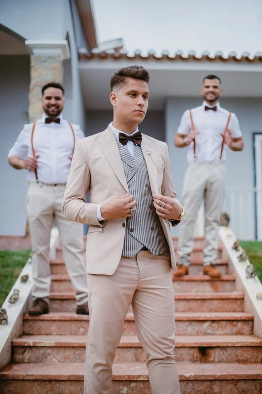 Three men dressed in formal attire, standing on outdoor stairs in front of a house. The man in the foreground wears a beige suit with a striped vest and bow tie, looking serious. The two men in the background wear white shirts with suspenders, bow ties, and light-colored pants, smiling and carrying backpacks.