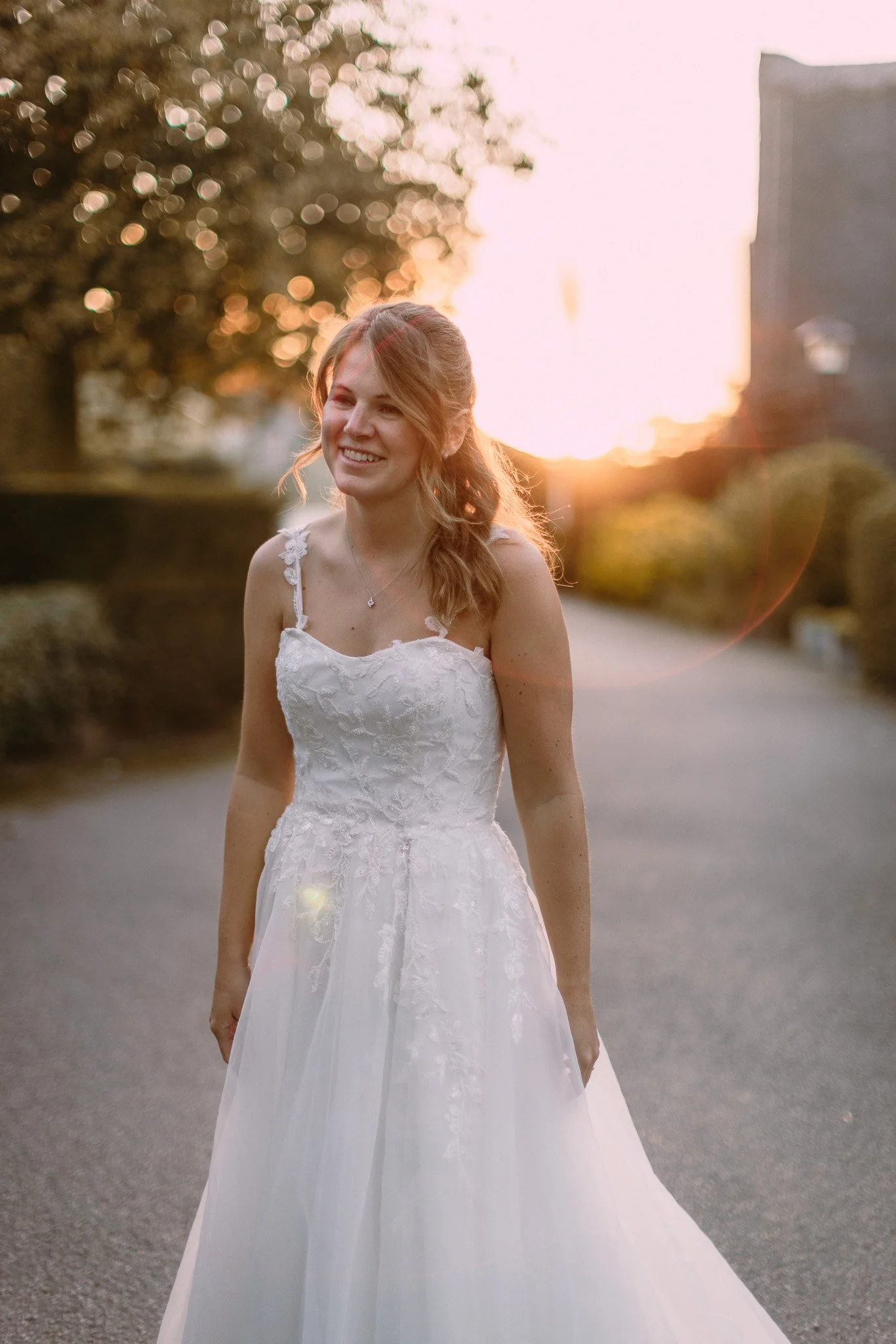 A woman in a white wedding dress standing outdoors at sunset, smiling.