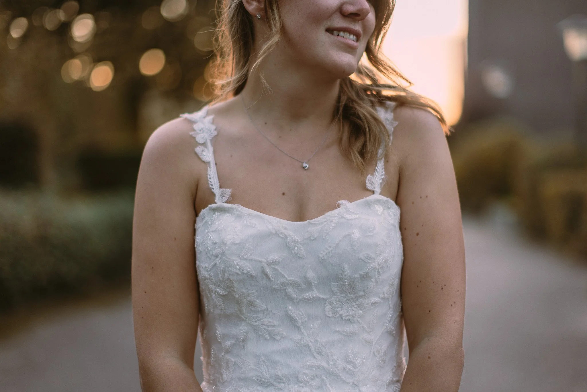 Close-up of a woman in a white lace dress, smiling and wearing a necklace, outdoors during sunset or golden hour.
